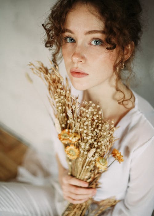 High angle of crop charming female with curly hair sitting with bouquet of dry plants and looking at camera