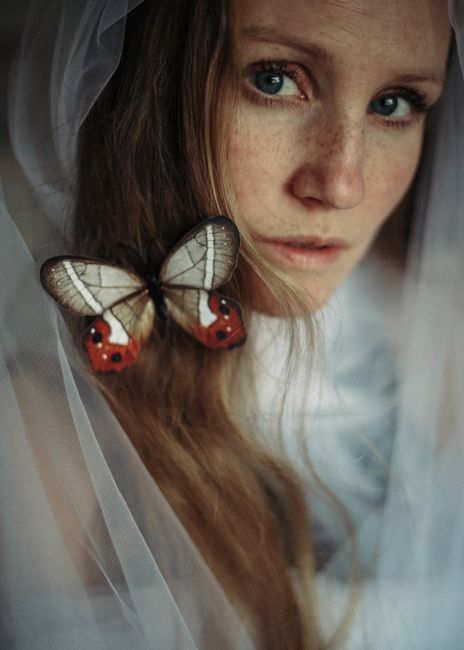 Crop serious female with freckles on face wrapped in thin veil with butterfly on long hair looking at camera
