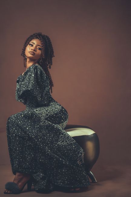 Charming African American woman with long curly hair in stylish long dress sitting on chair in studio against brown background and looking away