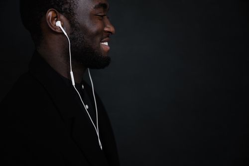 Free Side view of portrait of African American smiling male in black jacket with white earphones listening to music against black background Stock Photo