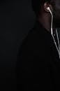 Side view of unrecognizable African American male in black jacket with white wired headphones standing against black background