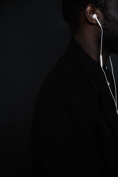Free Side view of unrecognizable African American male in black jacket with white wired headphones standing against black background Stock Photo