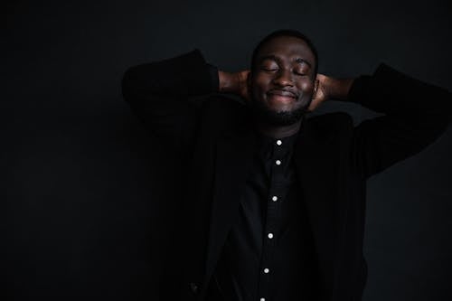 Cheerful young African American male in trendy black shirt with white buttons and black jacket with hands behind head on black background with closed eyes