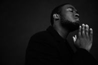 Monochrome handsome unshaven African American male in black suit praying with hands clasped under chin
