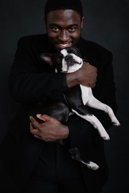 Good looking black male in black apparel on black background holding adorable Boston Terrier with eyes closed with both hands while smiling and positively looking at camera