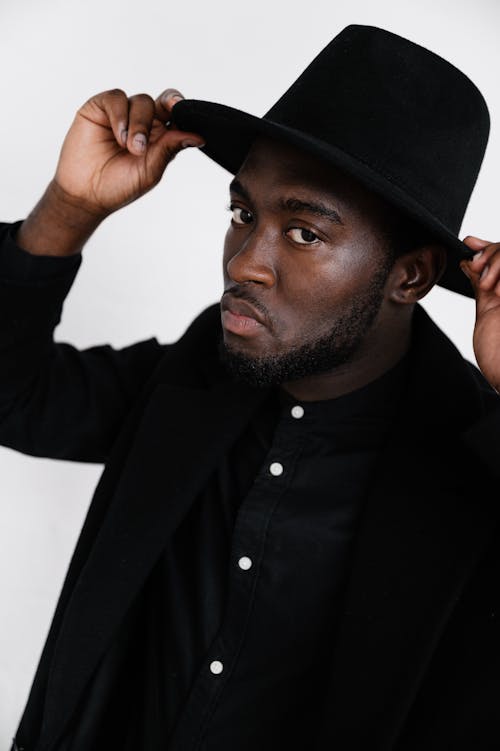 Serious adult black male wearing stylish black jacket and shirt touching hat fields with fingers while looking at camera