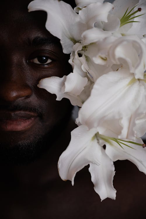 Crop African American male with bare shoulder holding delicate white lily flowers near face and looking at camera