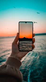 A hand holds a smartphone displaying a vibrant ocean sunset with birds flying overhead.