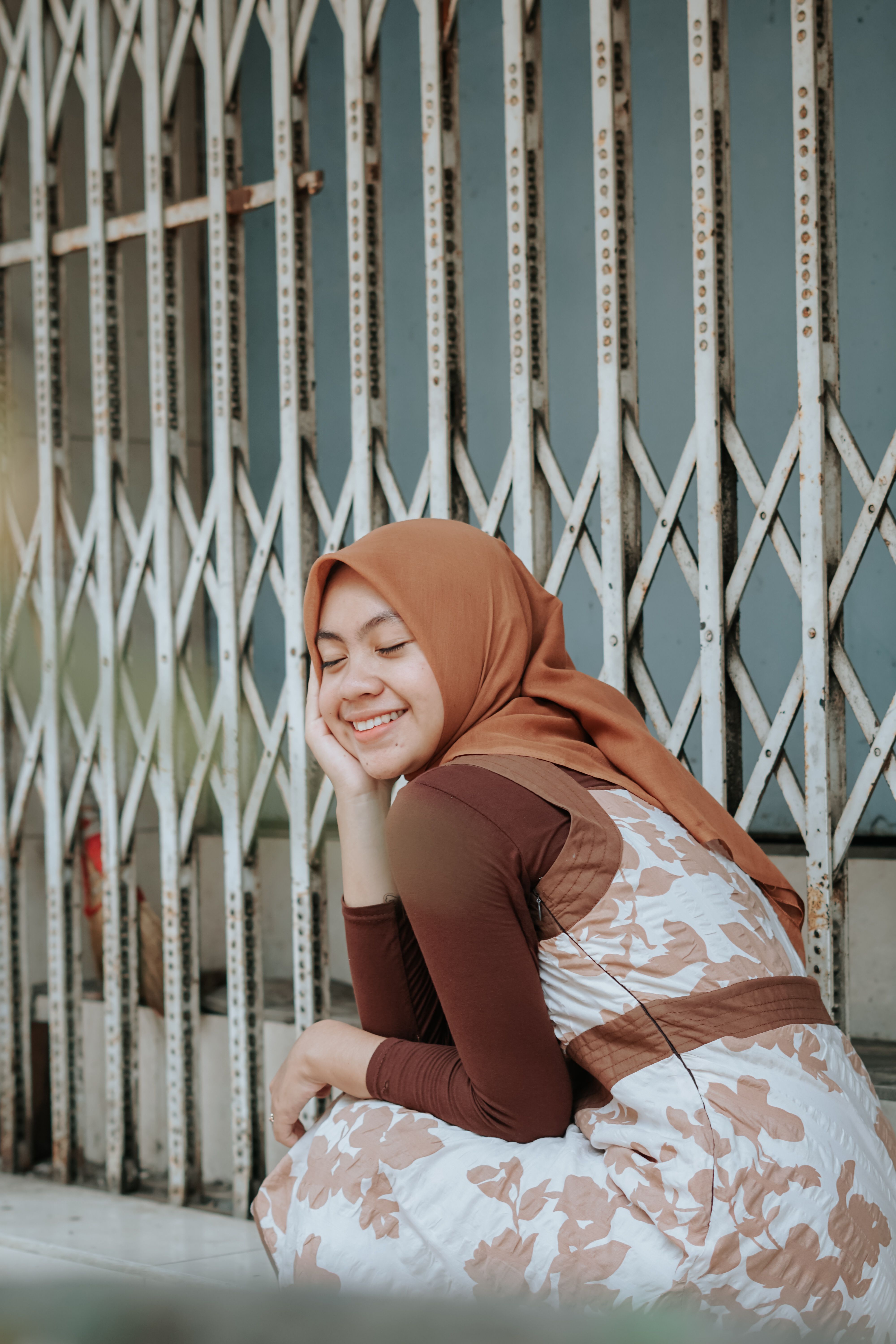 A joyful woman in a hijab sits with eyes closed against a metal folding gate, enjoying a serene moment outdoors.
