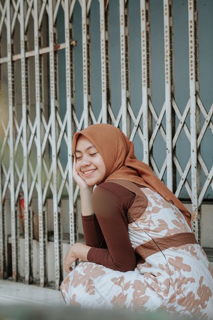 A joyful woman in a hijab sits with eyes closed against a metal folding gate, enjoying a serene moment outdoors.
