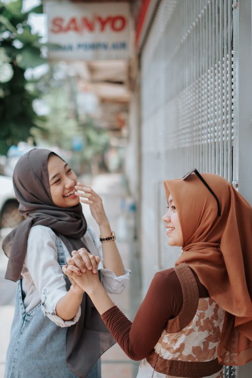 Free Two women in hijabs laughing and holding hands by a street gate on a sunny day. Stock Photo