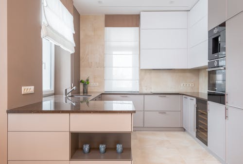 Free Empty contemporary kitchen with beige walls and light floor tile furnished with minimalist cupboards with gray panels equipped with oven and built in appliances Stock Photo