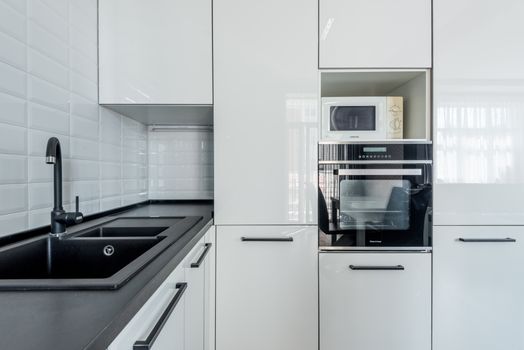 Interior of light black and white kitchen corner with white cupboards and black sink near white tiled wall and oven with microwave