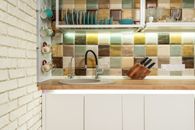 Interior of bright kitchen corner furnished with wooden counter and white cupboards decorated with various dishware and glasses against tiled wall
