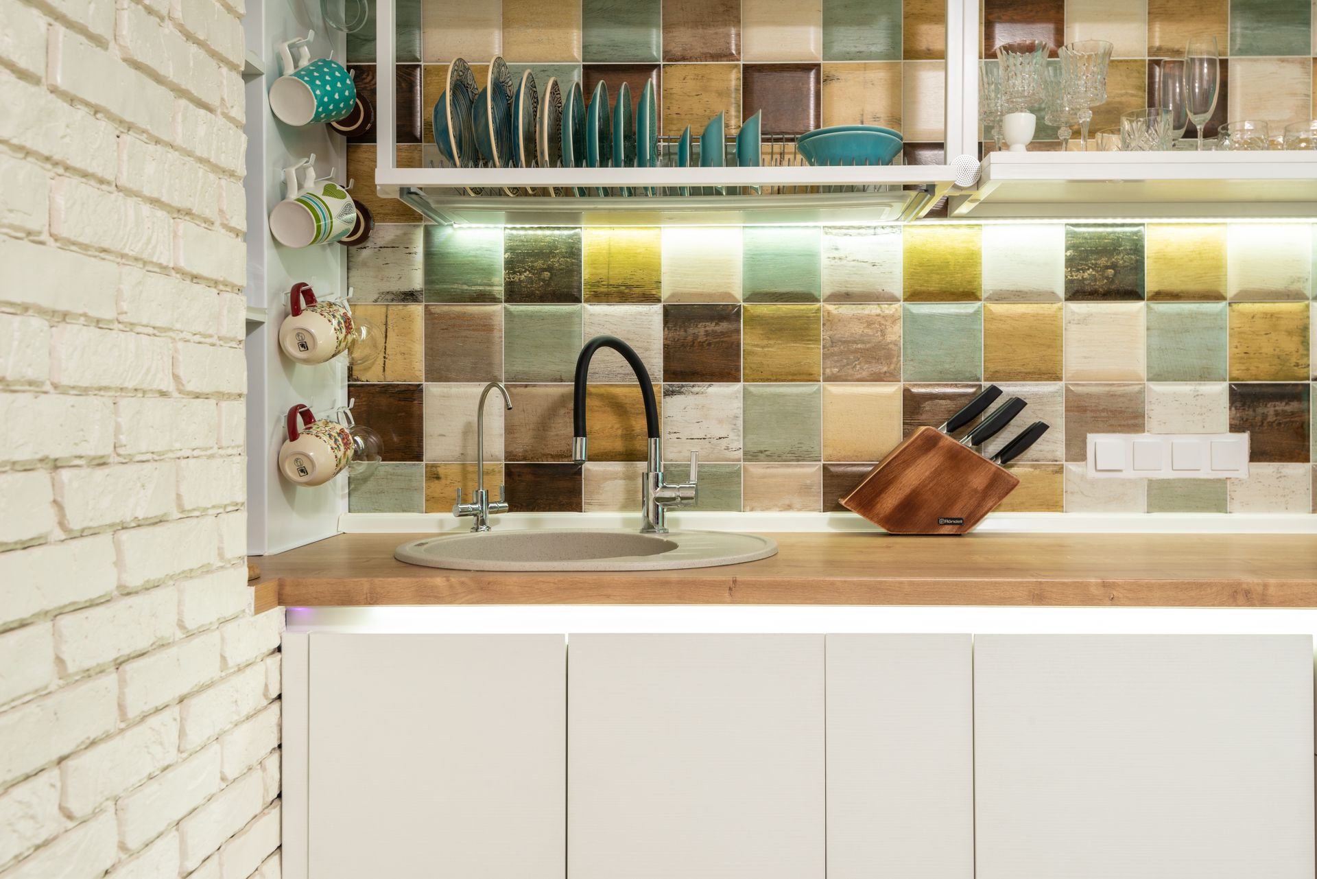 Interior of bright kitchen corner furnished with wooden counter and white cupboards decorated with various dishware and glasses against tiled wall