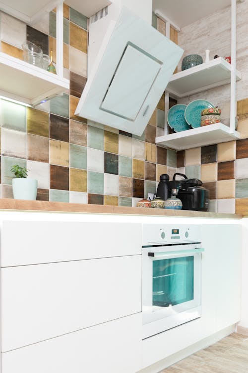 Interior of light kitchen furnished with white cupboards and shelves with oven decorated with various dishware with multicolored square shaped tiled wall