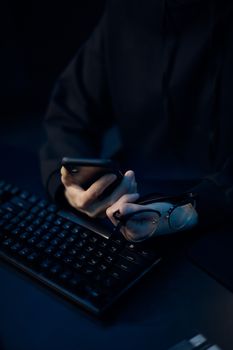 Close-up of hands holding phone and glasses over keyboard in dark setting.