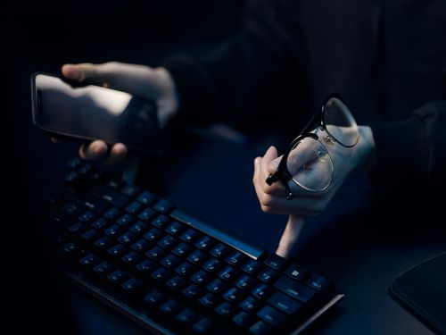 Free Close-up of hands using smartphone and computer keyboard, holding eyeglasses, focused workspace. Stock Photo
