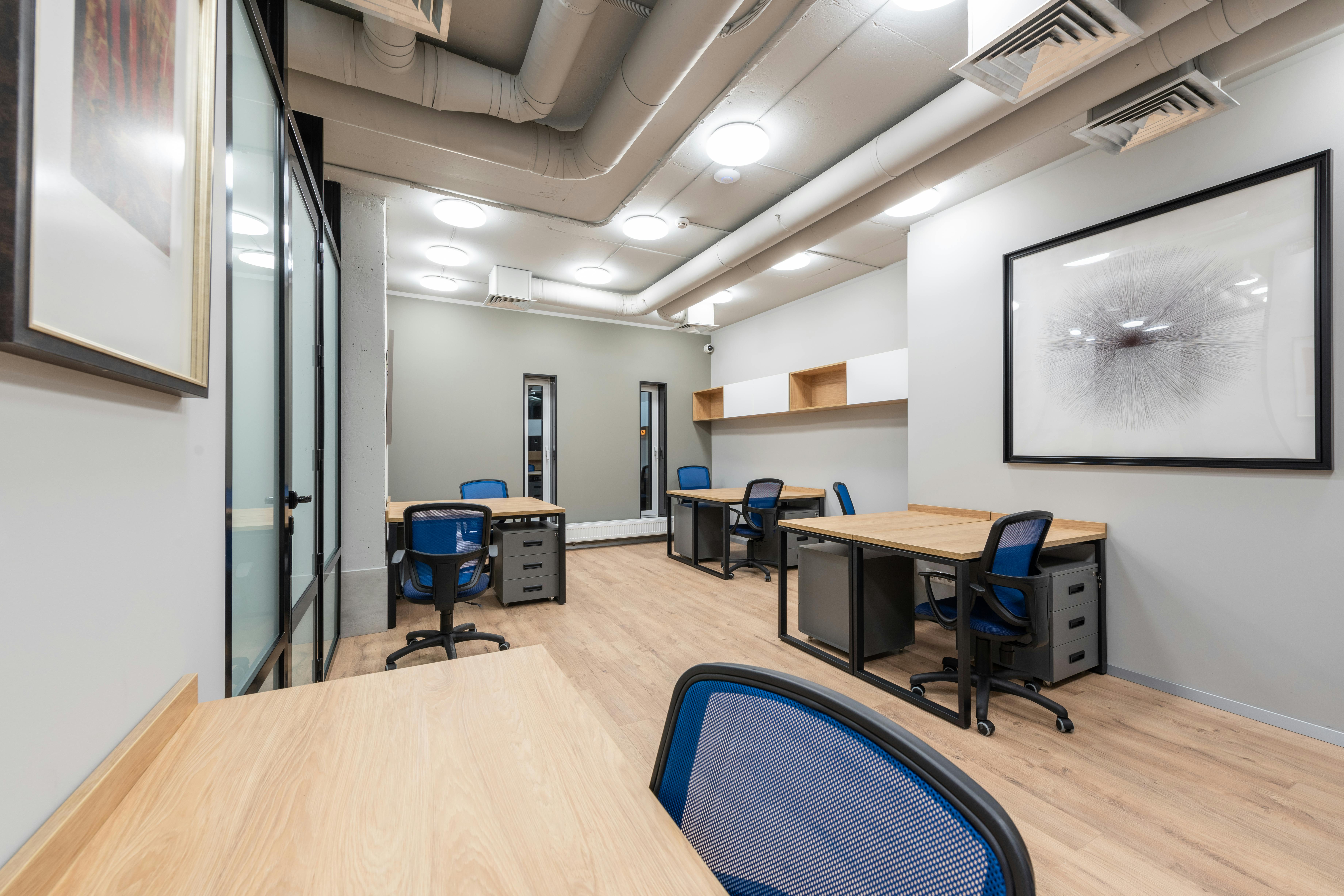 Modern office with wooden desks, blue chairs, and exposed ceiling ducts in a clean, minimal design.