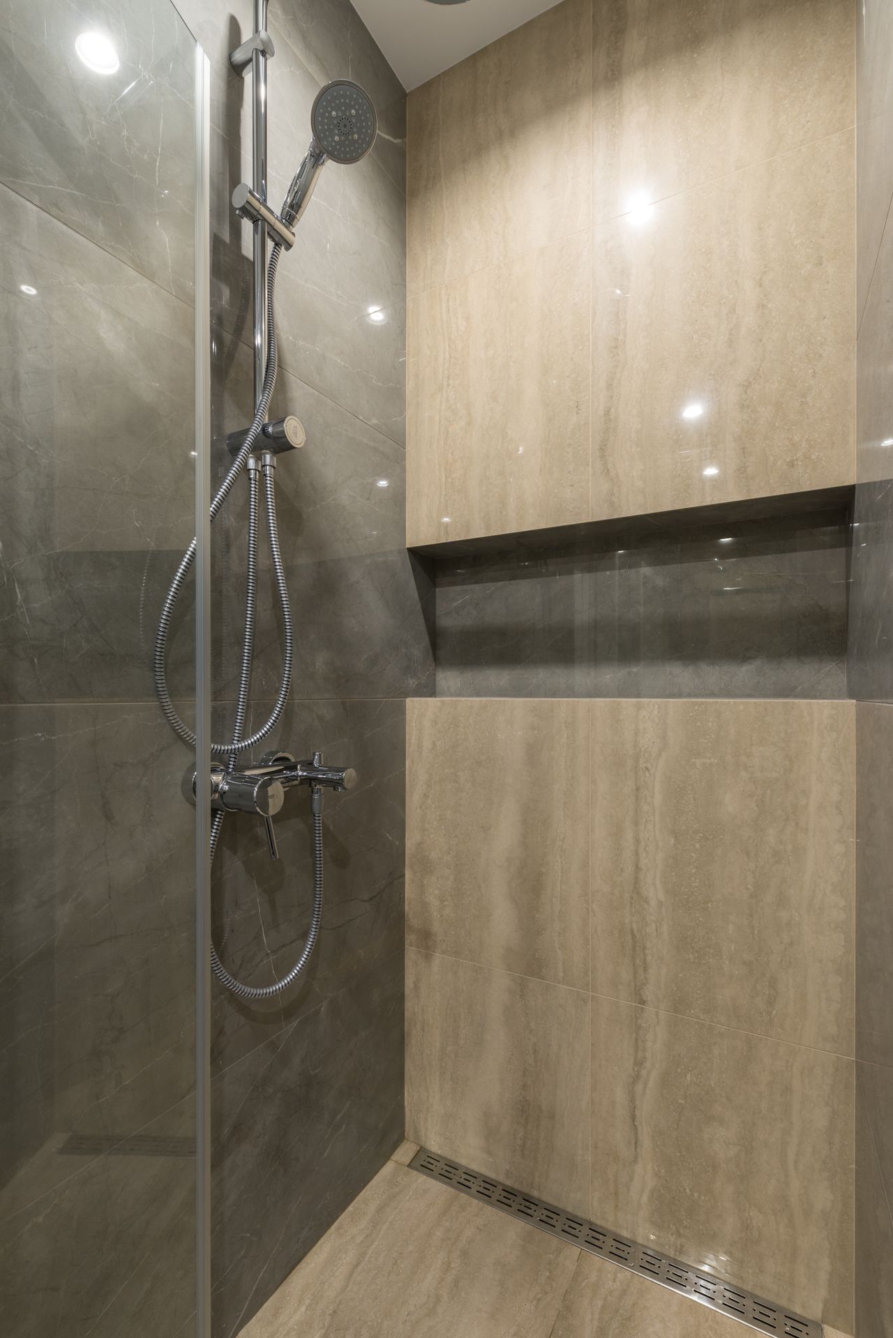 A grey marble shower interior with a handheld shower, tiled shelf niche and a linear floor drain