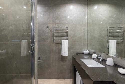 Interior of modern minimalist bathroom with glass shower cabin and shiny tiled walls reflecting in big mirror hanging above sink