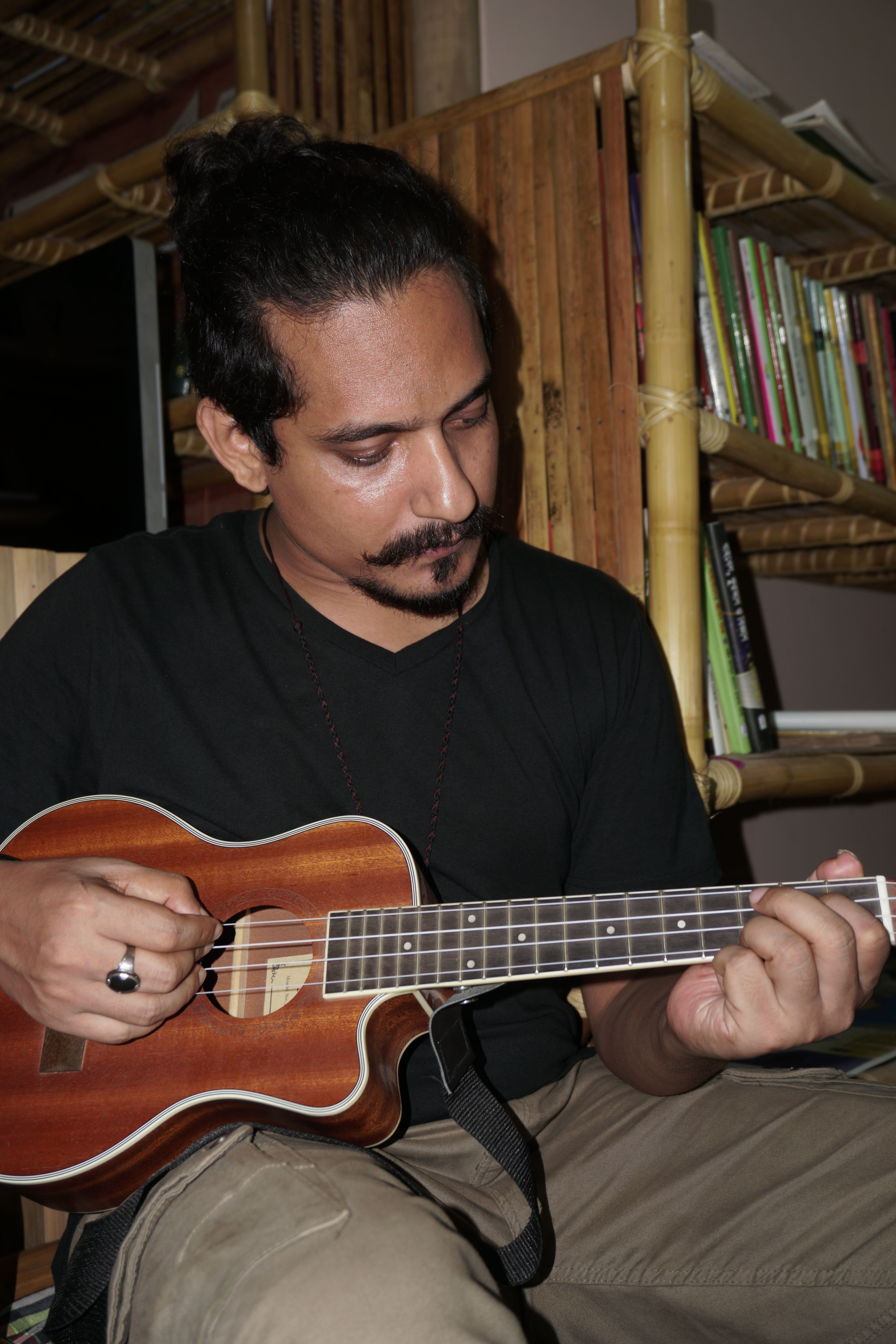 Man with facial hair playing ukulele indoors surrounded by books.