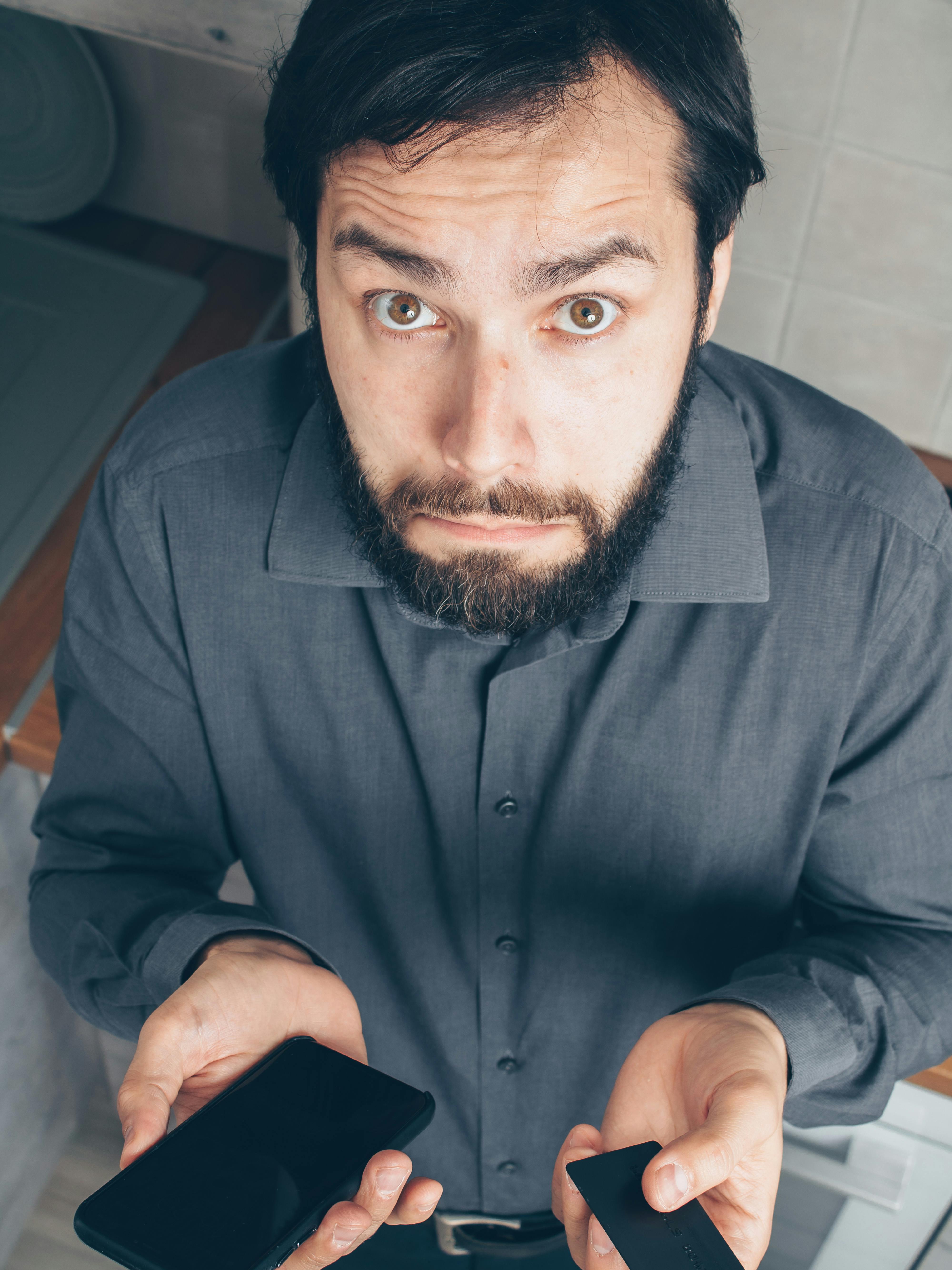 Bearded man in gray shirt looking confused while holding a smartphone and credit card.