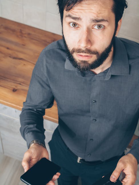 A perplexed man in a gray shirt holding a credit card and smartphone indoors.