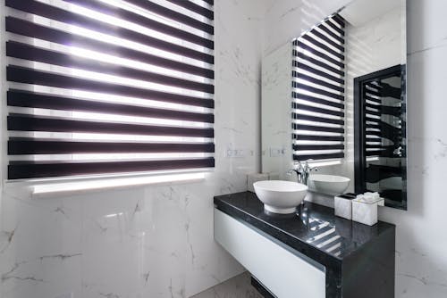 White sink and marble walls in modern bathroom with big mirror reflecting window with jalousie in daylight