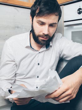 A worried man sitting indoors examining financial papers, depicting stress and concern.