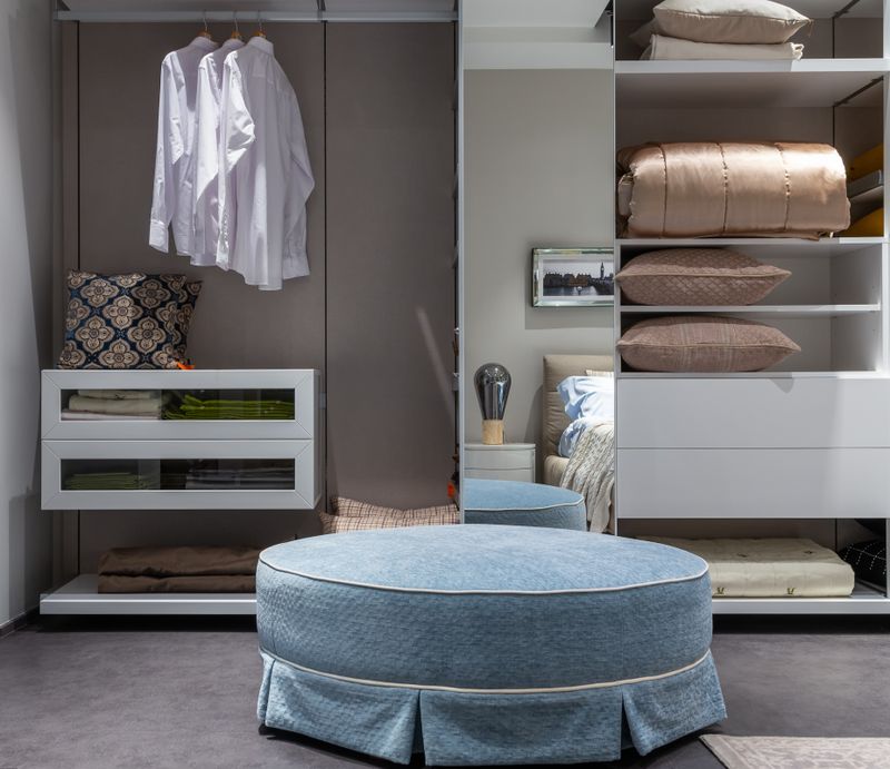 Round pouf beside an open wardrobe with neatly hung clothes and folded bedding