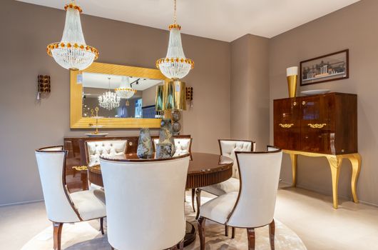 Chairs at table with decorative vases under glowing chandeliers placed near wall with and mirror and vintage styled cabinet in apartment