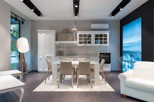 Chairs at table placed on rug near white cupboards and modern appliances at stylish kitchen with window and glowing lamp