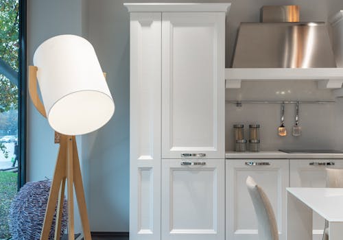 White cabinets and special utensils with extractor hood placed in modern kitchen with table and big shining stylish lamp near window