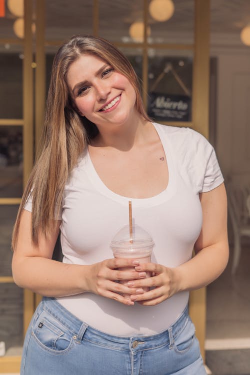 Woman in Denim Jeans Holding a Cup of Milkshake while Looking at the Camera