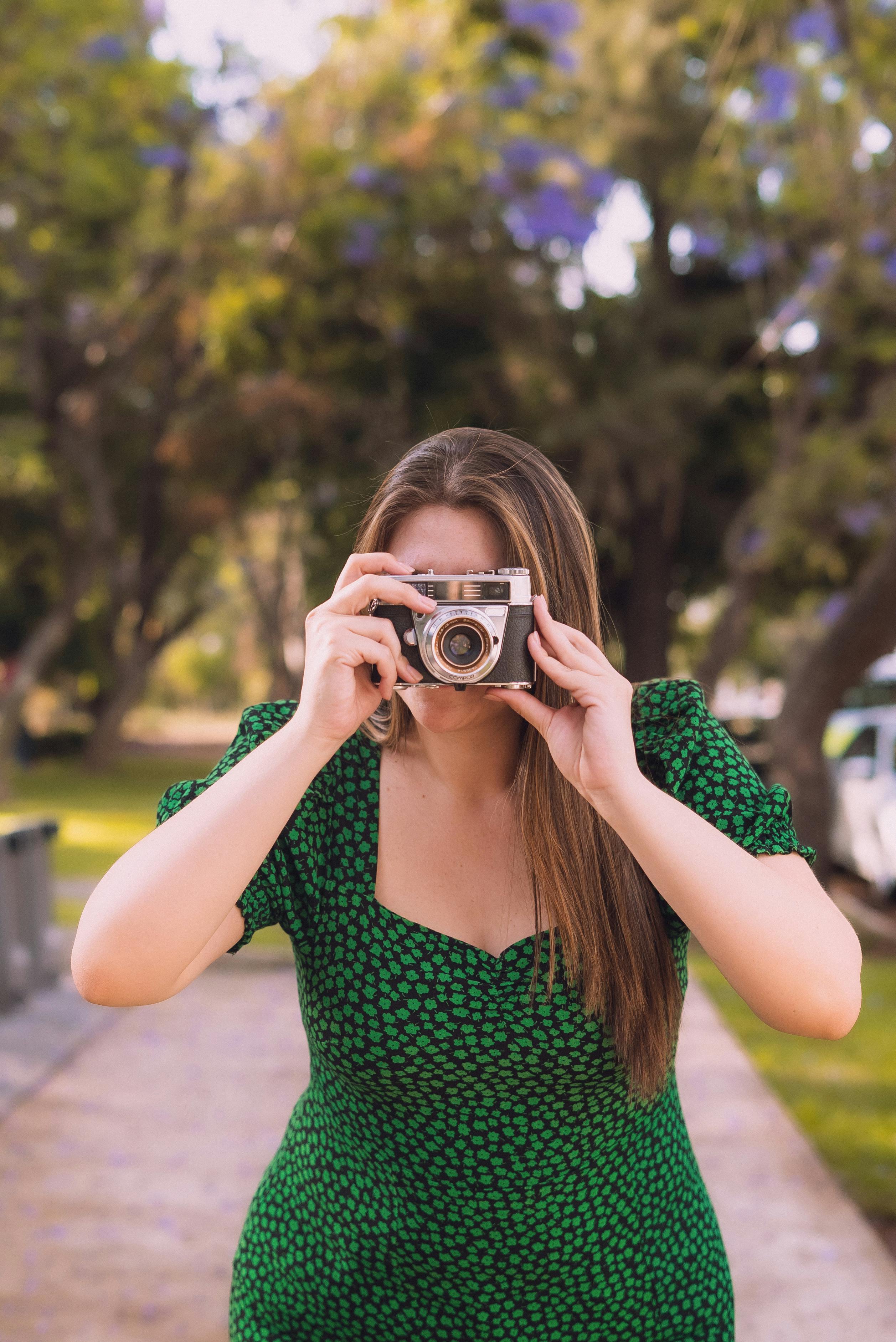 Woman in a green dress capturing a photo with a vintage camera outdoors during fall.