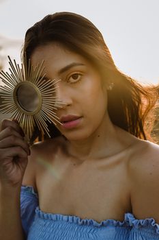 A woman in a blue off-shoulder top holding a brass sunburst mirror, captured in a dramatic portrait.