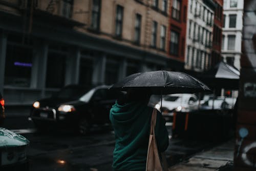 Back view of unrecognizable person in casual clothes walking with umbrella on city street in rainy day in evening time