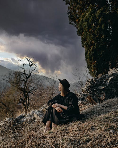 Female in hat contemplating mountain and leafless trees while sitting on dry grass and looking away in overcast weather