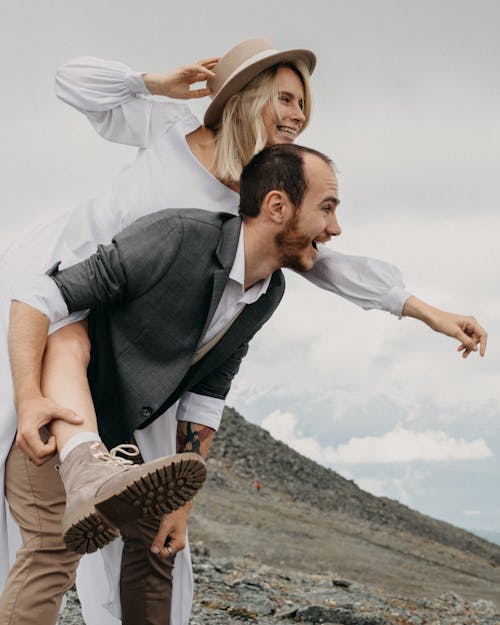 Cheerful trendy groom giving smiling bride piggyback ride while looking away on mount on wedding day
