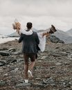 Anonymous trendy man carrying happy female beloved in white dress and boots while walking on mount with snow on wedding day