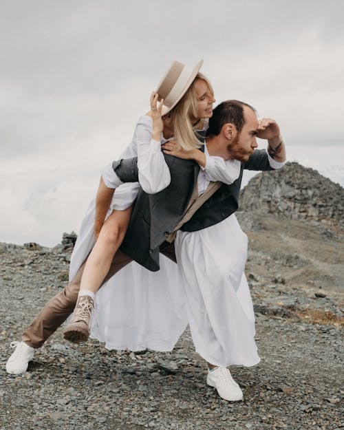 Trendy groom carrying smiling bride piggyback while looking into distance on mount under cloudy sky