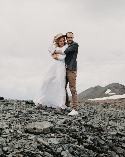 Free Trendy groom embracing bride in white dress while standing against mountain with snow on wedding day Stock Photo