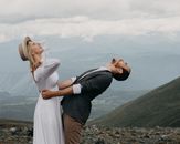 Side view of cheerful trendy groom embracing elegant bride in hat while looking up against mounts under cloudy sky