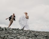 Full body of cheerful loving groom and bride in long white dress running on rough stones under cloudy gray sky