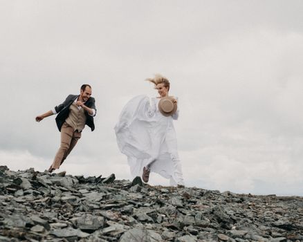 Full body of cheerful loving groom and bride in long white dress running on rough stones under cloudy gray sky