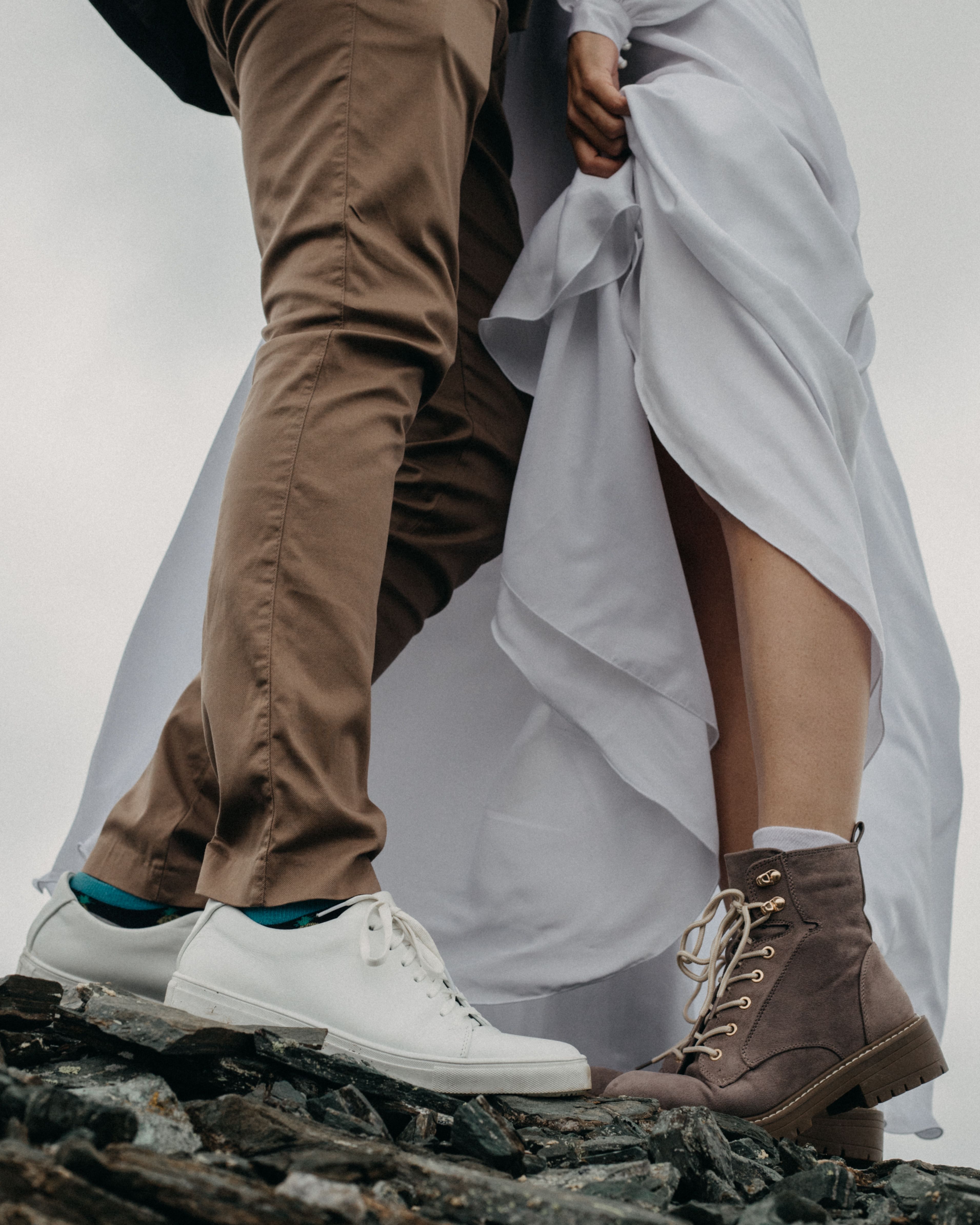 From below of crop unrecognizable female in long white dress and boots standing near boyfriend in brown suit and white sneakers on rocky ground in daytime
