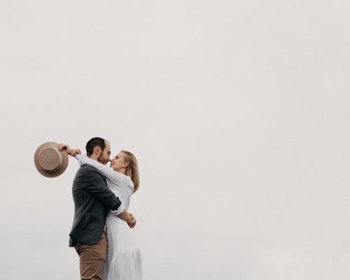 Free Side view of romantic man in stylish clothes standing and embracing female in white dress against cloudy sky in daytime Stock Photo