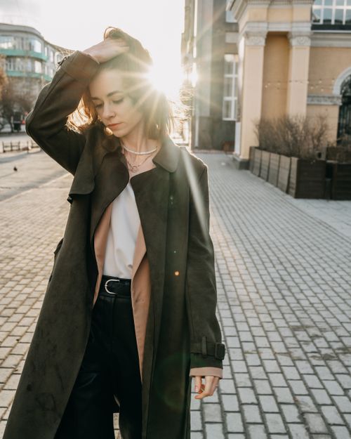 Free Young woman in stylish coat standing on city street and touching hair in sunny day Stock Photo
