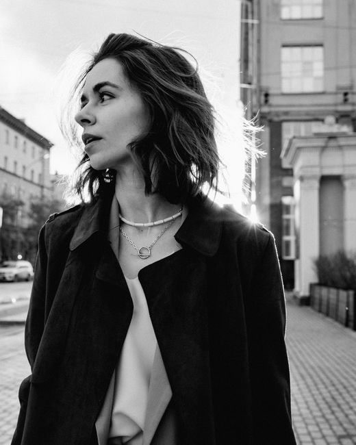 Black and white of pensive female with short hair in elegant coat standing on street and looking away in sunlight